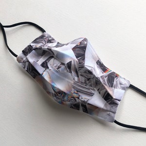 May include: A white face mask with a black elastic band. The mask has a geometric pattern of iridescent silver and black shapes.