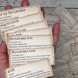 May include: A hand holds a stack of vintage-style Christmas-themed cards with definitions of words like "sugar plum", "Christmas Eve", and "gift". The cards are printed on aged paper with a typewriter font.