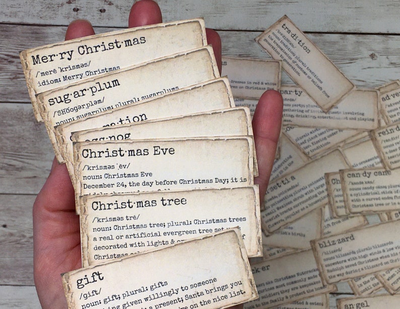 May include: A hand holds a stack of vintage-style Christmas-themed cards with definitions of words like "sugar plum", "Christmas Eve", and "gift". The cards are printed on aged paper with a typewriter font.