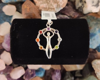 May include: Silver pendant featuring a stylized human figure with arms raised, surrounded by a circle of seven colorful gemstones. The pendant is displayed on a black velvet background.