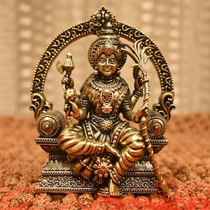 May include: A detailed bronze-colored statue of a seated deity, adorned with intricate jewelry and a crown. The statue holds various symbolic objects and is set against a blurred background. The statue is approximately 6 inches tall.