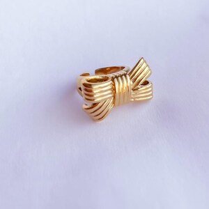 May include: A gold-toned ring with a bow design. The bow is made of multiple layers of metal, creating a textured look.