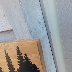 May include: Wooden sign with a natural wood grain finish. The sign features a black silhouette of pine trees. The sign is rectangular and has a beveled edge. The background includes white and gray siding.