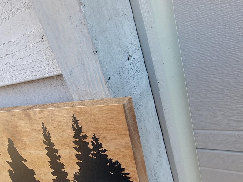 May include: Wooden sign with a natural wood grain finish. The sign features a black silhouette of pine trees. The sign is rectangular and has a beveled edge. The background includes white and gray siding.