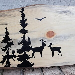 May include: A wooden sign featuring a black silhouette of a woodland scene. The scene depicts trees, deer, and a bird in flight. The wood has a natural, rustic appearance.