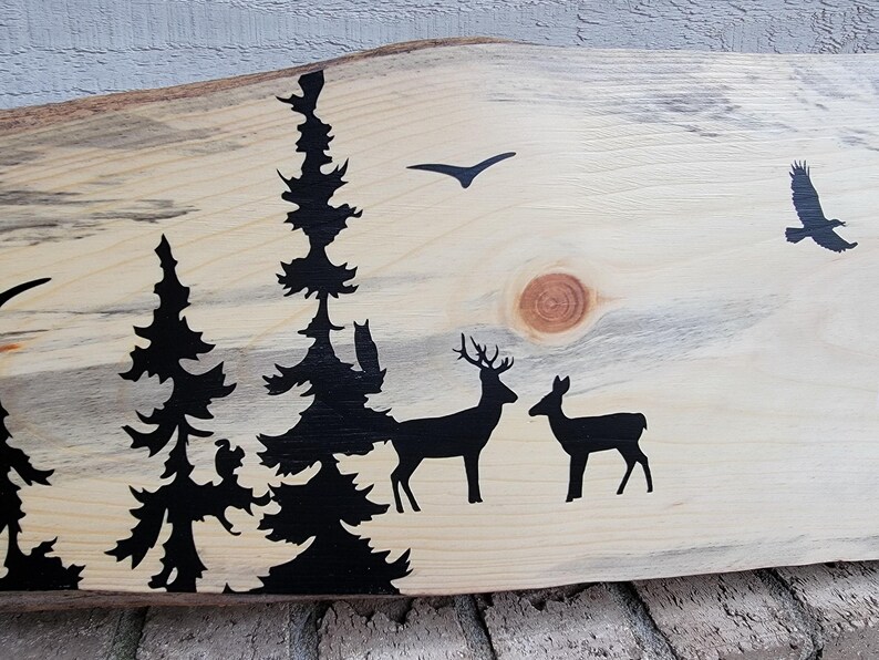May include: A wooden sign featuring a black silhouette of a woodland scene. The scene depicts trees, deer, and a bird in flight. The wood has a natural, rustic appearance.
