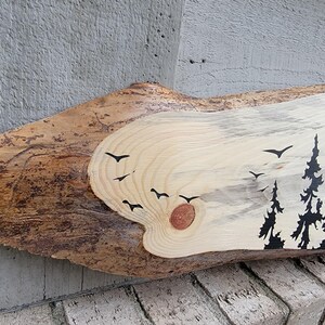 May include: A wooden slice with a natural wood grain pattern and a painted silhouette of a forest scene with trees and birds. The wood is light brown with darker brown streaks and a knot in the centre.