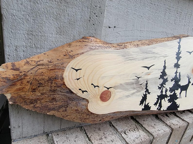 May include: A wooden slice with a natural wood grain pattern and a painted silhouette of a forest scene with trees and birds. The wood is light brown with darker brown streaks and a knot in the centre.
