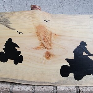 May include: A wooden sign featuring two black silhouettes of people riding quad bikes. The sign has a natural wood grain and a rough edge.