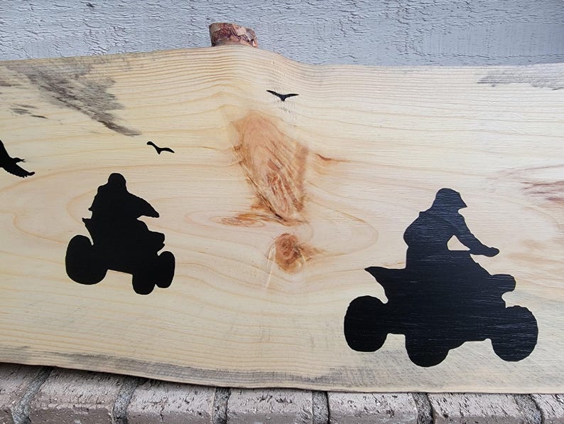 May include: A wooden sign featuring two black silhouettes of people riding quad bikes. The sign has a natural wood grain and a rough edge.