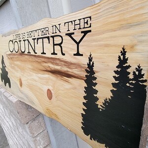 May include: Wooden sign with the text "Life is better in the country" in black lettering. The sign has a natural wood grain and features black silhouettes of pine trees.