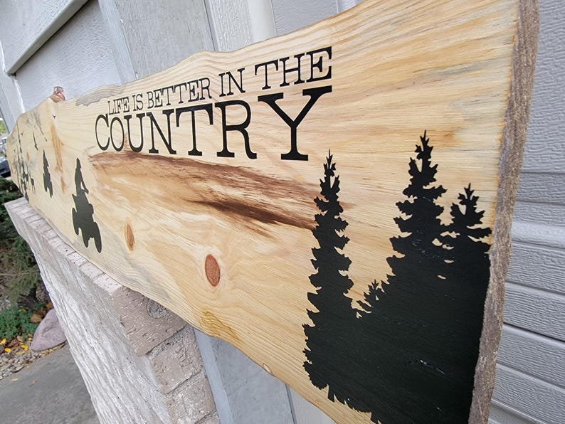 May include: Wooden sign with the text "Life is better in the country" in black lettering. The sign has a natural wood grain and features black silhouettes of pine trees.