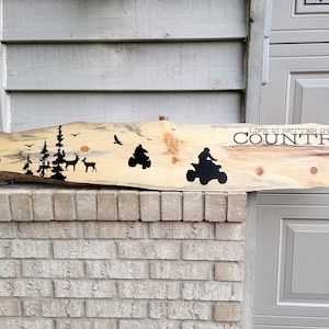 May include: A wooden sign with the text "Life is better in the country" painted in black. The sign also features silhouettes of trees, deer, and a quad bike.