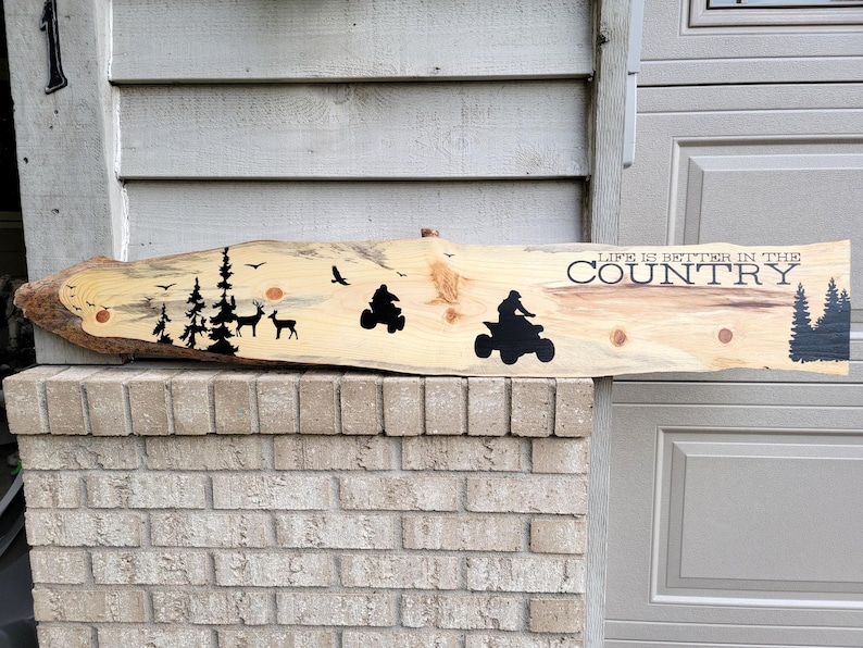 May include: A wooden sign with the text "Life is better in the country" painted in black. The sign also features silhouettes of trees, deer, and a quad bike.