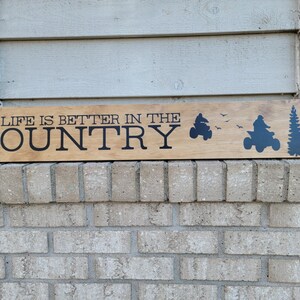 May include: Wooden sign with the phrase "LIFE IS BETTER IN THE COUNTRY" in black lettering. The sign features silhouettes of an ATV, birds, and trees. The sign is mounted on a brick wall.