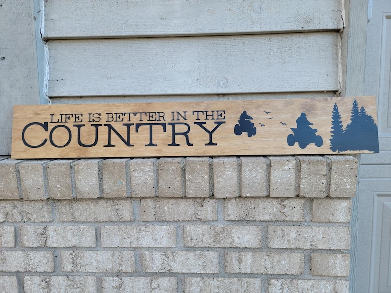 May include: Wooden sign with the phrase "LIFE IS BETTER IN THE COUNTRY" in black lettering. The sign features silhouettes of an ATV, birds, and trees. The sign is mounted on a brick wall.