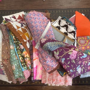 May include: A collection of various fabric pieces with diverse patterns and colors. Includes floral, geometric, and abstract designs in shades of pink, orange, green, and black. Some fabrics are rolled, others are folded, creating a visually rich assortment.