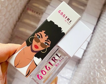 Puede incluir: Una botella de vidrio transparente con una etiqueta rosa que dice "GOGIRL By Nia". La botella está encima de una caja blanca con una ilustración en blanco y negro de una mujer con gafas de sol y un traje de baño blanco. La caja dice "GOGIRL By Nia".