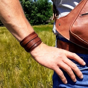 Può includere: Un braccialetto in pelle marrone con più cinturini. Il braccialetto è indossato al polso di una persona che indossa una camicia grigio chiaro e jeans blu.