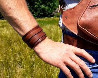 May include: A brown leather cuff bracelet with multiple straps. The bracelet is worn on the wrist of a person wearing a light gray shirt and blue jeans.