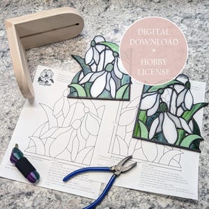 May include: A wooden display stand and two stained glass snowdrop flower panels with green and white details. A printed pattern, pliers, and a wrapped tool are also present. The image includes the text "DIGITAL DOWNLOAD HOBBY LICENSE".