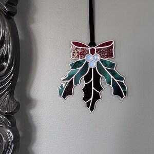 K&ouml;nnte beinhalten: Ein Weihnachtsornament aus Glas in Form eines Stechpalmenblattes mit einer roten Schleife. Das Blatt ist gr&uuml;n und schwarz mit drei blauen Glasbeeren. Das Ornament h&auml;ngt an einem schwarzen Band.