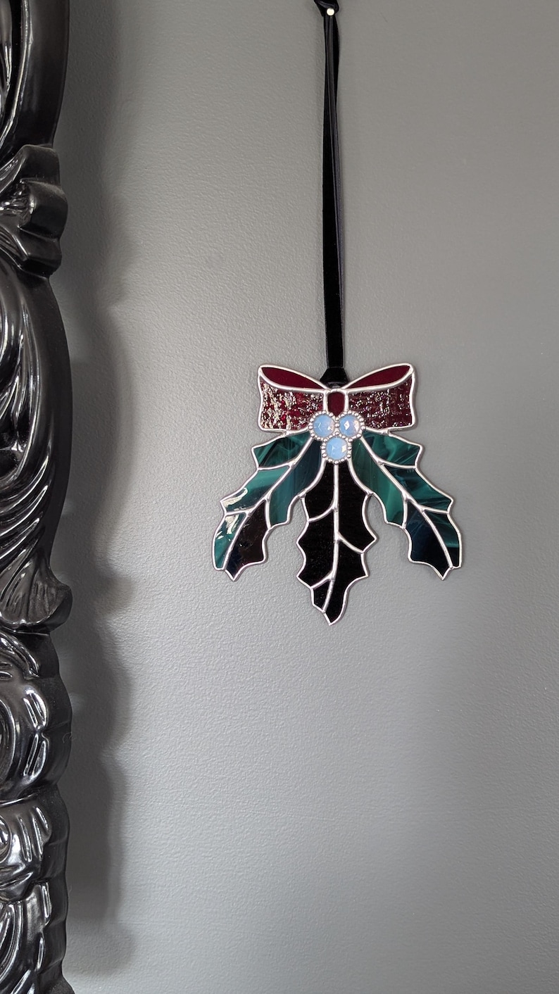 K&ouml;nnte beinhalten: Ein Weihnachtsornament aus Glas in Form eines Stechpalmenblattes mit einer roten Schleife. Das Blatt ist gr&uuml;n und schwarz mit drei blauen Glasbeeren. Das Ornament h&auml;ngt an einem schwarzen Band.