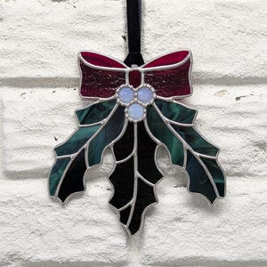 K&ouml;nnte beinhalten: Weihnachtsschmuck aus Glas in Form eines Stechpalmenblattes mit roter Schleife, gr&uuml;nen Bl&auml;ttern und drei hellblauen Beeren. Der Schmuck ist mit Silber verziert und h&auml;ngt an einem schwarzen Band. Eine festliche Weihnachtsdekoration.