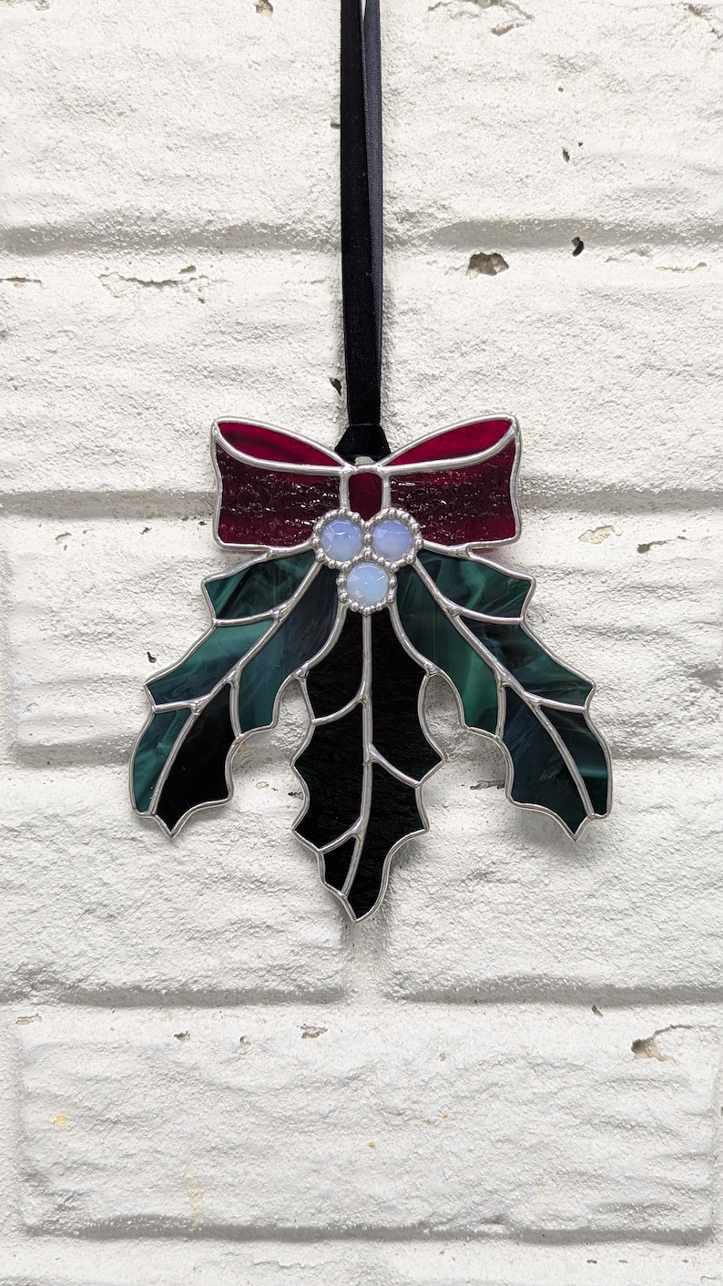 K&ouml;nnte beinhalten: Weihnachtsschmuck aus Glas in Form eines Stechpalmenblattes mit roter Schleife, gr&uuml;nen Bl&auml;ttern und drei hellblauen Beeren. Der Schmuck ist mit Silber verziert und h&auml;ngt an einem schwarzen Band. Eine festliche Weihnachtsdekoration.