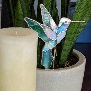 May include: A stained glass hummingbird garden stake in shades of blue, green, and white, with a silver border. The hummingbird is placed in a potted plant next to a cream-colored candle.
