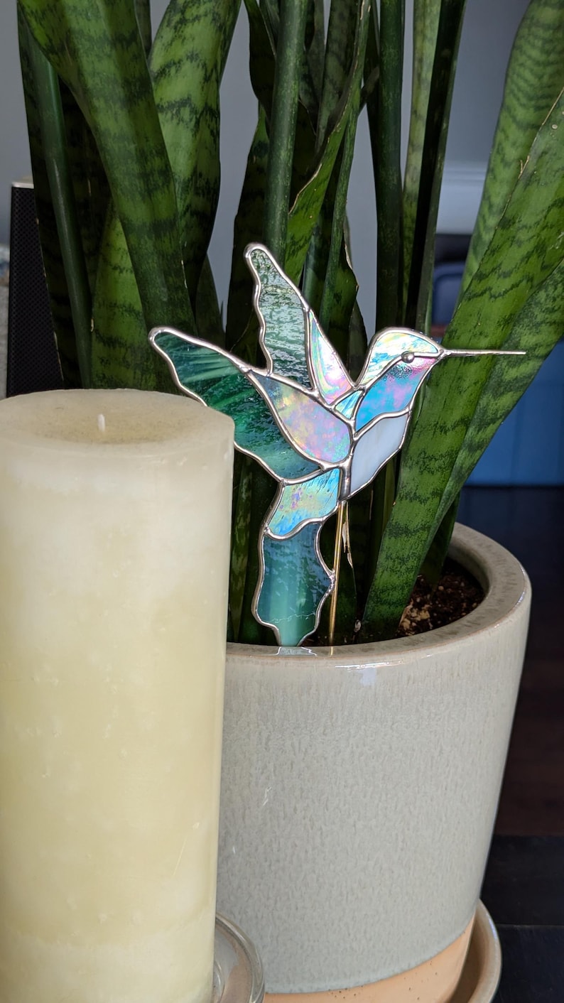 May include: A stained glass hummingbird garden stake in shades of blue, green, and white, with a silver border. The hummingbird is placed in a potted plant next to a cream-colored candle.