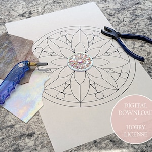 Op de afbeelding: Een mandala-patroon van glas-in-lood met een middelste stuk van iriserend glas. Het patroon is afgedrukt op een wit vel papier. Er liggen ook stukken glas, een glassnijder en een tang op de tafel.