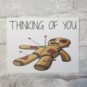 May include: A white card with the text "Thinking of You" printed in black. A cartoon voodoo doll with red stitches and pins is pictured below the text.