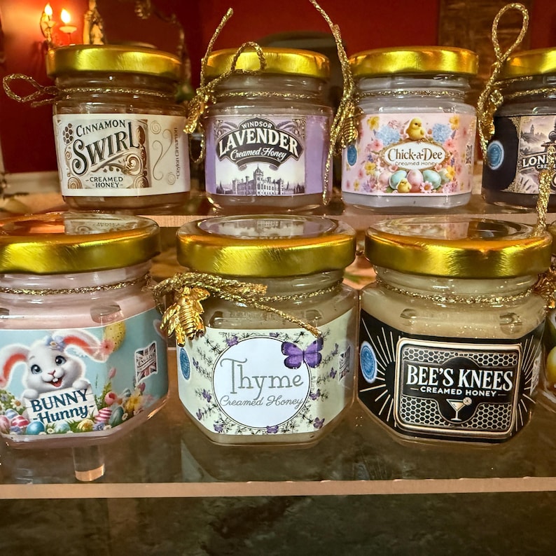 May include: A collection of small glass jars with gold lids, filled with creamed honey. Each jar has a decorative label with text such as "Cinnamon Swirl", "Lavender", "Chick-a-Dee", "Bunny Hunny", "Thyme", and "Bee's Knees".