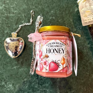 May include: A jar of strawberry creamed honey with a gold lid and a decorative label featuring strawberries, bees, and flowers. A heart-shaped tea infuser and a decorative spoon are next to the jar. A pink ribbon is tied around the jar.
