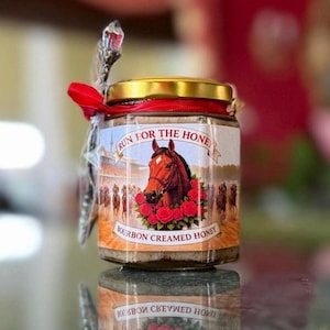 May include: A jar of bourbon creamed honey with a gold lid and a red ribbon. The label features a horse head surrounded by red roses and the text "RUN FOR THE HONE". A decorative spoon is attached to the jar.