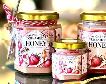 May include: Three glass jars of strawberry creamed honey with gold lids. The labels feature strawberries, bees, and the text "Strawberry Creamed Honey." The jars are tied with red and white twine.