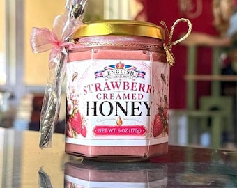 May include: A hexagonal glass jar of strawberry creamed honey with a gold lid, featuring a label that reads English Creamed Honey Strawberry Honey Net Wt. 6 oz (170g). A small silver spoon is attached to the side with a pink ribbon and gold cord.