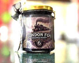 Könnte beinhalten: Ein Glas London Fog Cremehonig mit goldenem Deckel und einem Etikett mit einer Vintage-Zug-Illustration. Das Etikett enthält den Text "London Fog Creamed Honey" und die Nummer "6". Um das Glas ist ein schwarzes Band gebunden.