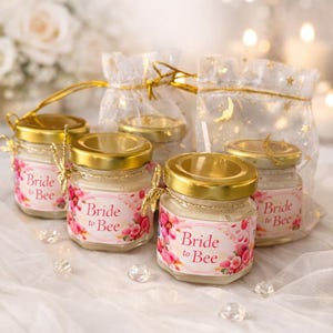 May include: Small glass jars with gold lids, labeled "Bride to Bee" with floral accents. The jars are arranged with small organza bags, all on a white surface. Decorative crystals are scattered around the jars.