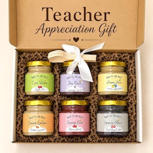 May include: A gift box labeled "Teacher Appreciation Gift" contains six small jars with gold lids and labels. The labels read "Core Values," "Zen Desk," "Power Hour," "Sweet Escape," "Energy Kick," and "Steamy Memo." The box includes wooden spoons and a white ribbon.
