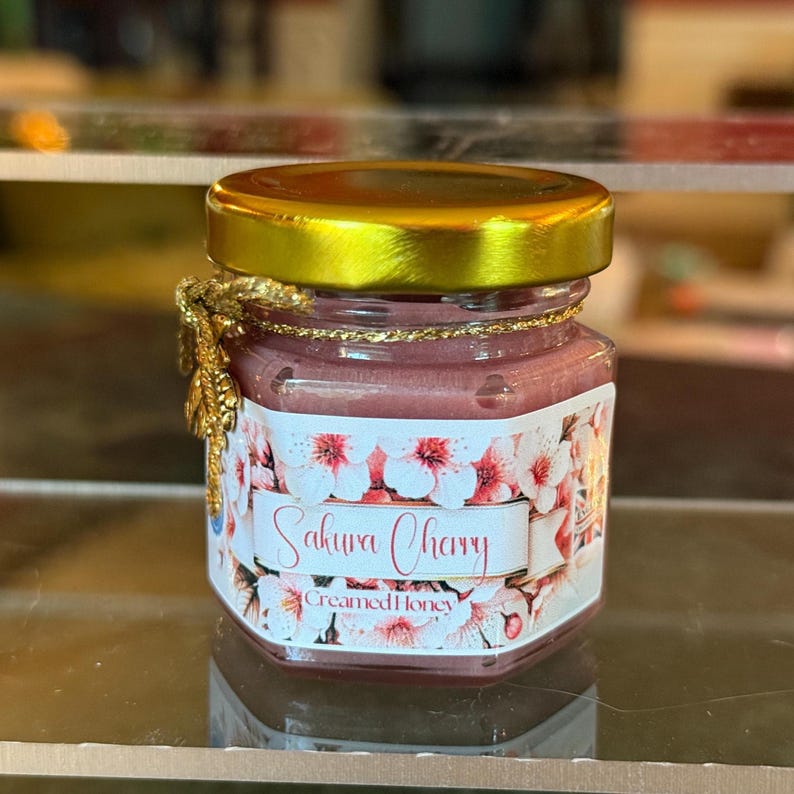 May include: A small, hexagonal glass jar filled with pink-hued creamed honey. The jar has a gold-colored lid and a label with the words "Sakura Cherry" and "Creamed Honey" over a floral design. A gold-colored decorative element is attached to the jar.