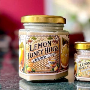 May include: A glass jar of creamed honey with a gold lid, featuring a label that reads Lemon Honey Hugs Creamed Honey, decorated with illustrations of bees, a lemon slice, and a teacup, alongside a smaller matching jar of creamed honey.
