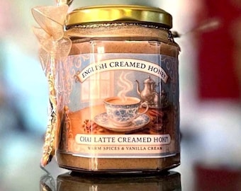 May include: A jar of English Creamed Honey with a gold lid and a label featuring a teacup and teapot. The label reads "Chai Latte Creamed Honey" with "Warm Spices & Vanilla Cream" below. A decorative spoon and bow are attached.