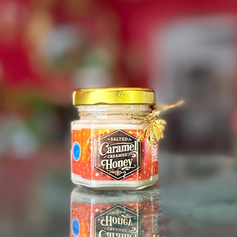 May include: A small glass jar of salted caramel creamed honey with a gold lid and a decorative gold bee. The label reads "Salted Caramel Creamed Honey" in an ornate font. The jar is reflected in the surface below.