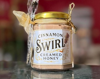 May include: A jar of Cinnamon Swirl Creamed Honey with a gold lid and label. The label features the words "Cinnamon Swirl Creamed Honey" in elegant script. A small spoon is tied to the jar with a gold ribbon.