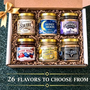 May include: A gift box filled with six small jars of flavored creamed honey. Flavors include Cinnamon Swirl, Honey Moon Vanilla, Windsor Lavender, Berry Berry, Lemon Honey Hugs, and Bee's Knees. The box is decorated with a gold bow and the text "26 FLAVORS TO CHOOSE FROM".