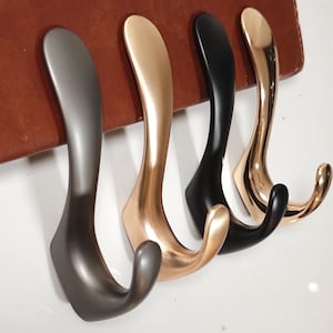 Op de afbeelding: Vier decoratieve metalen wandhaken op een rij, elk met een gebogen ontwerp. De haken zijn in tinten grijs, goud, zwart en brons. Ze zijn gemonteerd op een bruin leerachtig oppervlak tegen een witte achtergrond.