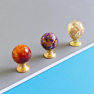 Könnte beinhalten: Drei dekorative Schrankknöpfe mit Messingbasen. Einer ist rot und orange, ein anderer lila und gold, und der dritte ist transparent mit Muscheln im Inneren. Diese Knöpfe verleihen jedem Möbelstück eine einzigartige Note.