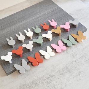 Könnte beinhalten: Eine Sammlung dekorativer Schrankknöpfe in verschiedenen Formen und Farben. Die Knöpfe umfassen Hasen-, Mickey Mouse- und Schmetterlingsdesigns in Weiß-, Grau-, Rosa-, Orange-, Grün- und Goldtönen. Sie sind auf einer grauen Holzoberfläche angeordnet.